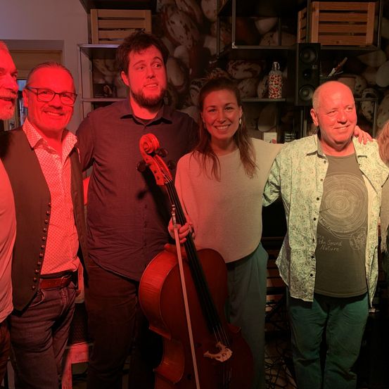 Fünf Menschen lächeln und posieren für ein Foto in einem Raum. Ein Mann mit Bart hält ein Cello. Hinter ihnen sind ein Lautsprecher und Regale mit verschiedenen Gegenständen zu sehen.