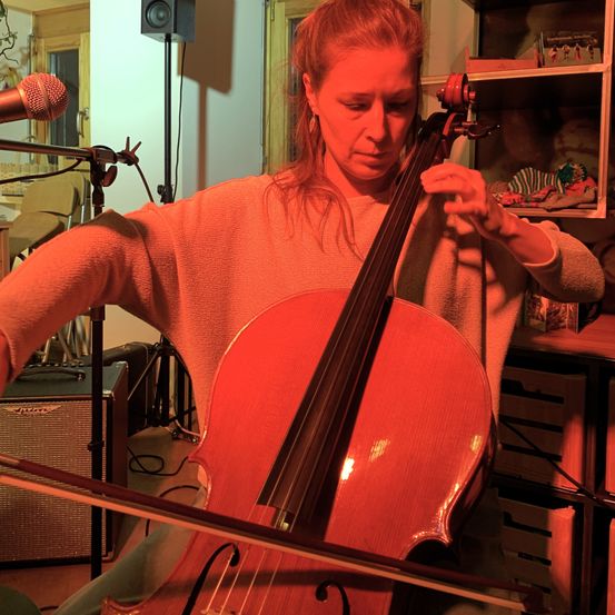 Eine Frau mit roten Haaren spielt Cello, beleuchtet von rotem Licht. Ein Mikrofon steht vor ihr.
