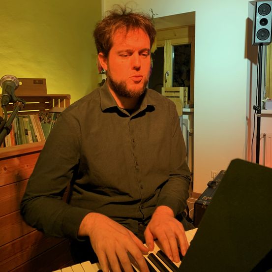 Ein Mann sitzt an einem Klavier und spielt es, während er nach unten schaut. Hinter ihm steht ein Mikrofon auf einem Ständer und ein Lautsprecher. Bücher sind auf einem Regal links von ihm gestapelt, und ein Fenster mit Blick auf ein Gebäude draußen befindet sich rechts von ihm.