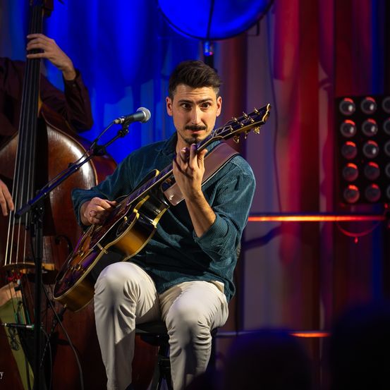 Ein Mann spielt eine E-Gitarre auf der Bühne, mit einem Bassisten hinter ihm und einem Mikrofon davor. Die Bühne ist mit blauen und roten Lichtern beleuchtet.