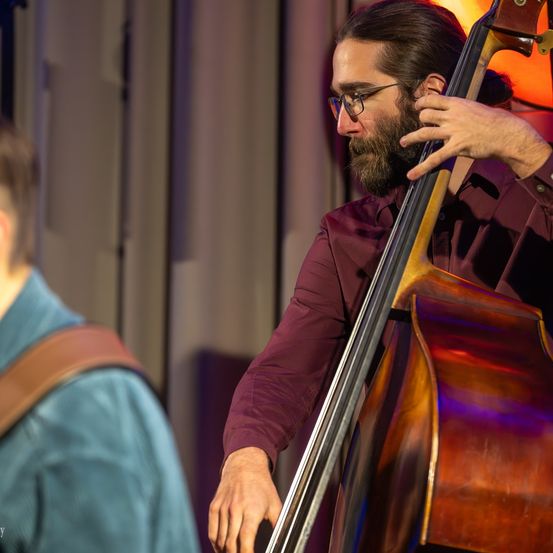 Ein Mann spielt auf der Bühne den Kontrabass, trägt eine Brille und ein weinrotes Hemd, während ein anderer Mann in einem blauen Hemd im Hintergrund steht.