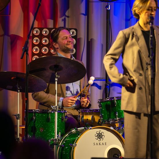 Ein Mann spielt auf der Bühne Schlagzeug mit Becken, während eine Frau neben ihm in ein Mikrofon singt.