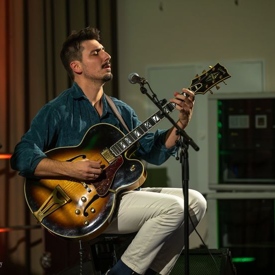 Ein Mann spielt eine Gitarre auf der Bühne, sitzt auf einem Hocker mit einem Mikrofon vor ihm. Er trägt ein türkisfarbenes Hemd und weiße Hose. Hinter ihm ist ein Vorhang und ein Fenster.