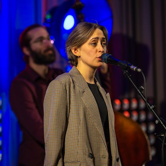 Eine Frau singt mit ernster Miene in ein Mikrofon. Hinter ihr spielt ein Mann Cello. Die Bühne ist mit blauen und roten Lichtern beleuchtet.