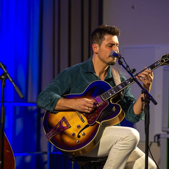 Ein Mann tritt auf der Bühne auf, spielt eine blaue und goldene E-Gitarre und singt in ein Mikrofon, während er auf einem Hocker vor blauen Vorhängen und Bühnenlichtern sitzt.