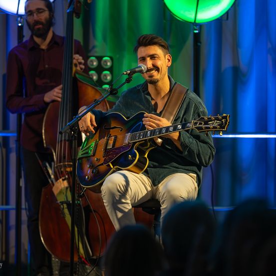 Ein Mann spielt Gitarre auf der Bühne und lächelt, während er für ein Live-Publikum auftritt. Hinter ihm spielt ein weiterer Musiker Cello, und ein grünes Licht strahlt auf die Bühne.