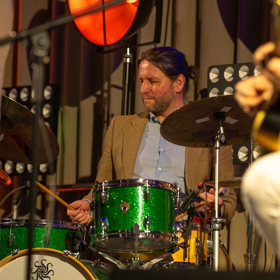 Ein Mann spielt eine Drum-Set mit grünen Trommeln und Becken, trägt ein blaues Hemd und einen braunen Blazer. Er sieht konzentriert aus, umgeben von verschiedenen Musikinstrumenten und Ausrüstung.
