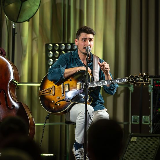 Ein Mann tritt auf der Bühne auf und spielt sitzend eine Gitarre. Er trägt ein grünes Hemd und weiße Hose. Hinter ihm steht ein Cello.