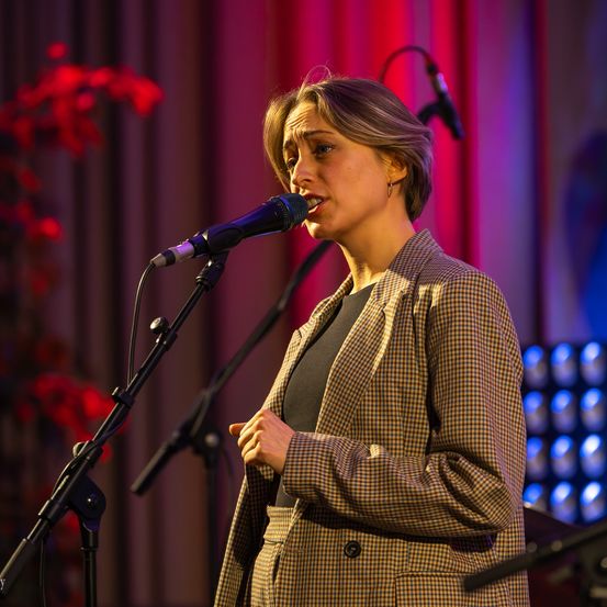 Eine Frau singt in ein Mikrofon auf der Bühne, mit einem roten Vorhang und Blumen im Hintergrund. Sie trägt einen karierten Blazer über einem schwarzen Oberteil.