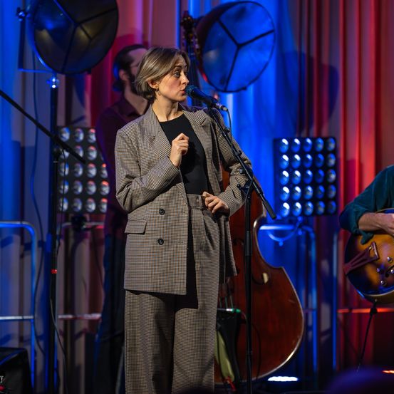 Eine Frau singt auf der Bühne, während ein Mann im Hintergrund Gitarre spielt. Die Bühne ist mit blauen und roten Vorhängen geschmückt.