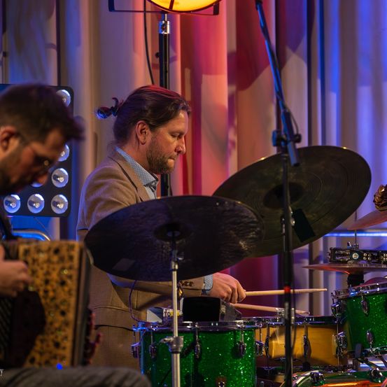 Zwei Musiker treten auf der Bühne auf. Einer spielt Akkordeon, der andere Schlagzeug und Becken. Der Schlagzeuger hat einen Pferdeschwanz. Scheinwerfer und ein Vorhang sind im Hintergrund.