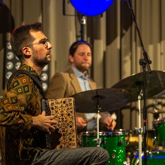 Ein Mann spielt Akkordeon, während ein anderer Mann Schlagzeug spielt. Die Szene spielt sich drinnen ab, mit einem weißen Vorhang im Hintergrund.