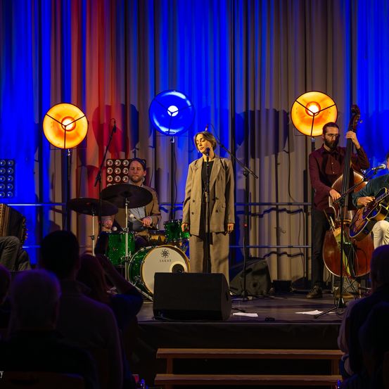 Eine Live-Band tritt auf der Bühne auf, mit einer Sängerin, einem Schlagzeuger und einem Cellisten. Das Publikum sitzt davor.