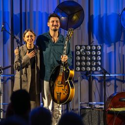 Ein Mann und eine Frau stehen lächelnd auf der Bühne, der Mann hält eine Gitarre. Mikrofone und ein Lautsprecher stehen dahinter. Das Publikum sitzt davor.