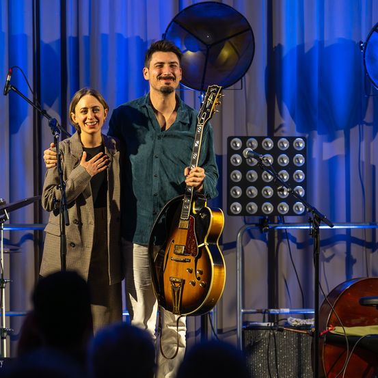 Ein Mann und eine Frau stehen lächelnd auf der Bühne, der Mann hält eine Gitarre. Mikrofone und ein Lautsprecher stehen dahinter. Das Publikum sitzt davor.