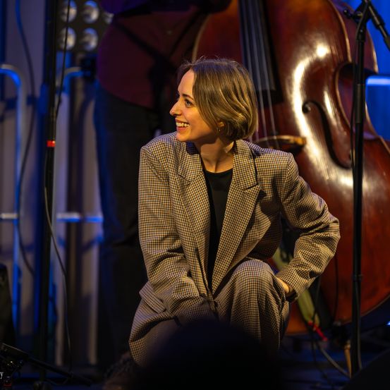 Eine Frau in einem karierten Anzug kniet lächelnd auf der Bühne, mit einem Cello und einem Mikrofonständer im Hintergrund. Die Bühne ist mit blauen Lichtern beleuchtet.