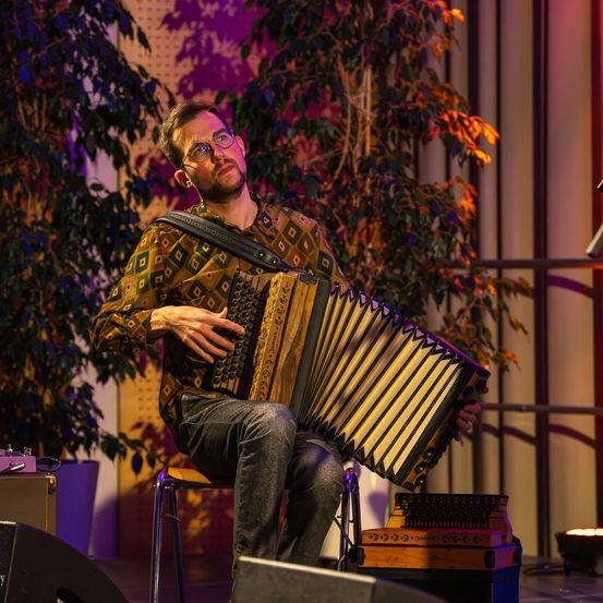 Ein Mann spielt Akkordeon auf der Bühne, sitzt auf einem Hocker, mit Pflanzen und Scheinwerfer im Hintergrund.