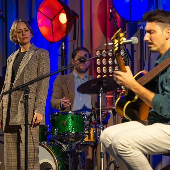 Eine Frau in einem Anzug steht auf der Bühne mit einer Band. Ein Mann spielt Gitarre, während ein anderer Mann Schlagzeug spielt. Ein rotes Licht beleuchtet die Bühne.