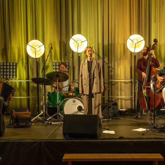 Eine Frau singt auf einer Bühne mit einem gelben Vorhang im Hintergrund. Hinter ihr spielen vier Männer Musikinstrumente. Es gibt zwei Schlagzeuge, einen Cello und ein Akkordeon.