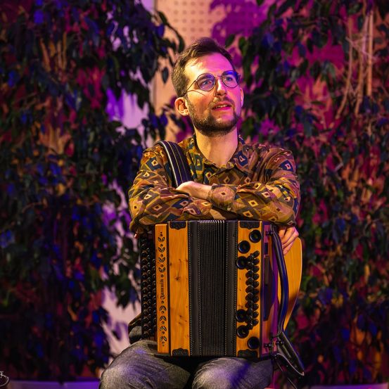 Ein Mann mit Brille sitzt auf einem Stuhl und hält ein Akkordeon, lächelt in die Kamera.