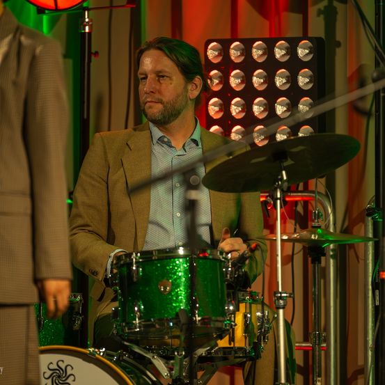 Ein Mann mit Bart und Schnurrbart spielt eine Schlagzeug-Set auf der Bühne, mit einem grünen Schlagzeug und einer Becken. Er trägt einen Anzug und ein Hemd.