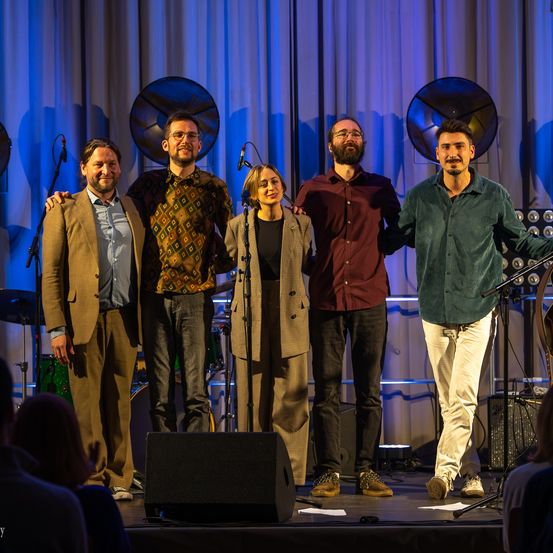 Fünf Personen stehen auf einer Bühne, mit Mikrofonen und Musikinstrumenten im Blickfeld. Drei Männer und zwei Frauen lächeln und posieren für ein Foto.