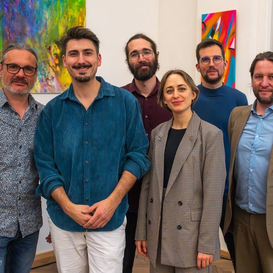 Sechs Personen, darunter fünf Männer und eine Frau, posieren für ein Foto in einer Galerie. Die Männer tragen Brillen und die Frau einen Blazer. Sie stehen vor einer weißen Wand, die mit farbenfrohen Gemälden geschmückt ist.