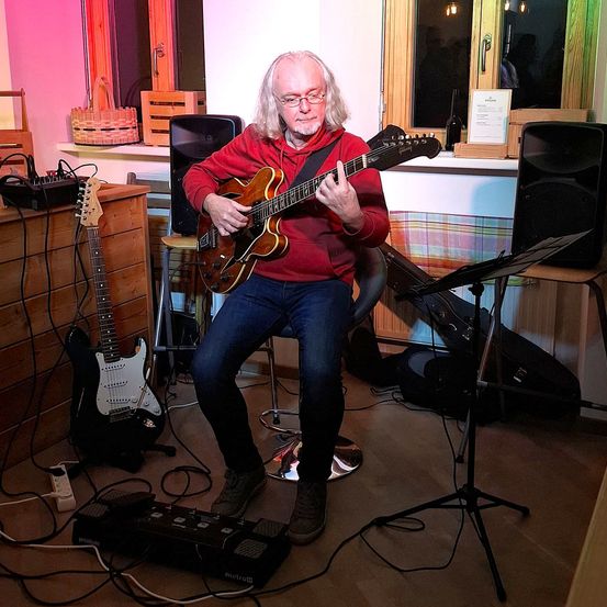Ein älterer Mann mit Brille spielt eine E-Gitarre in einem Raum mit rosa Wänden und Holzmöbeln. Eine schwarze Gitarre steht auf einem Ständer in der Nähe.