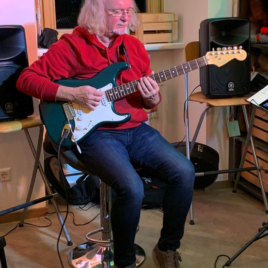 Ein älterer Mann mit langen Haaren und einer Brille sitzt auf einem Hocker und spielt eine E-Gitarre. Er trägt eine rote Kapuzenjacke und blaue Jeans. Hinter ihm stehen Lautsprecher und ein Holzschrank mit Lagerboxen.