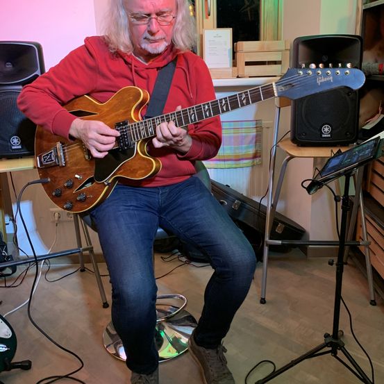 Ein älterer Mann mit langen Haaren, der eine rote Kapuzenjacke und Jeans trägt, sitzt auf einem Hocker und spielt eine E-Gitarre. Hinter ihm stehen Lautsprecher, ein Gitarrenkoffer und ein Holztisch.