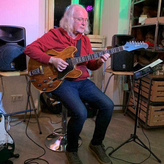 Ein älterer Mann mit langen Haaren und Brille spielt eine E-Gitarre, während er auf einem Hocker in einem Raum mit Musikequipment und Lagerboxen sitzt.