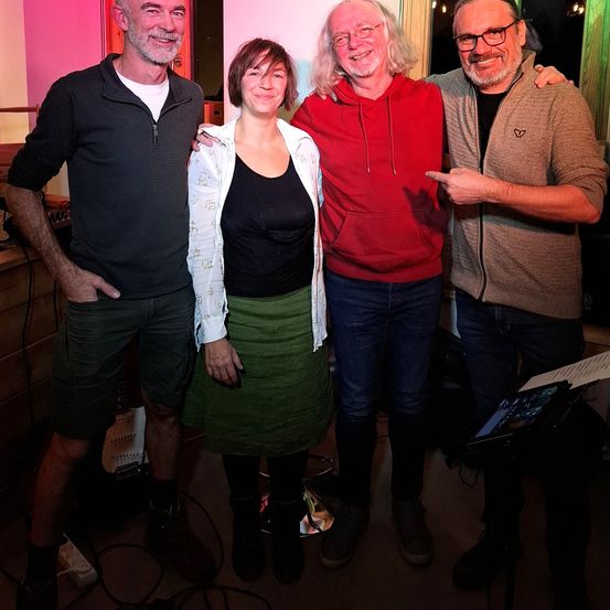 Vier Personen stehen gemeinsam in einem Raum und lächeln für ein Foto. Ein Mann in einem schwarzen Shirt und Shorts steht links, während eine Frau in einem grünen Rock und einer weißen Jacke in der Mitte ist. Neben ihr steht ein Mann in einer roten Kapuzenjacke und lächelt. Ein Mann in einer braunen Jacke und mit einer Brille steht rechts.