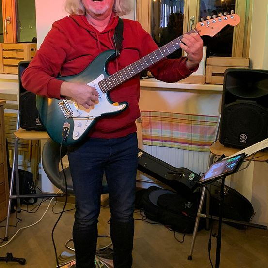 Ein Mann steht in einem Raum, lächelt und spielt eine E-Gitarre. Er trägt eine rote Kapuzenjacke und blaue Jeans. Hinter ihm sind ein Lautsprecher und ein Notenständer mit einem Tablet zu sehen.