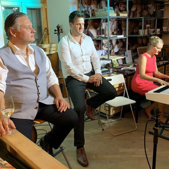 Drei Personen sind in einem Raum, zwei Männer und eine Frau. Die Frau spielt Klavier, während die Männer sitzen. Der Raum ist mit Büchern und Musikinstrumenten gefüllt.