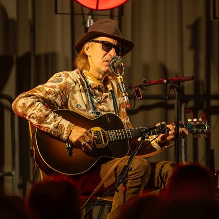 Ein Musiker in Hut und Sonnenbrille spielt Gitarre auf einer Bühne mit einem Mikrofon vor sich. Dahinter steht eine rote Lampe.