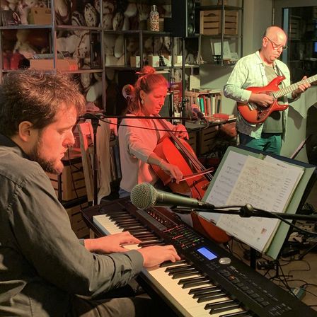 Ein Musiker spielt Klavier, während ein anderer Cello spielt und ein Dritter Gitarre. Sie sind in einem Raum mit Musikinstrumenten und Regalen mit Büchern und Gegenständen.