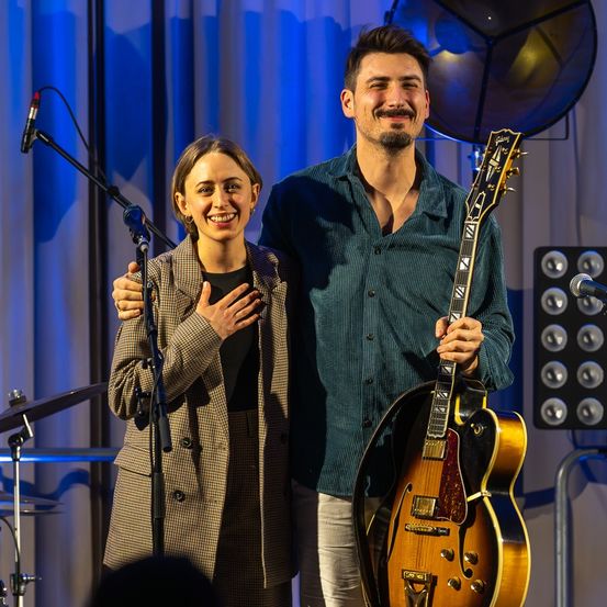Ein Mann und eine Frau stehen lächelnd auf der Bühne, der Mann hält eine Gitarre und die Frau umarmt ihn. Hinter ihnen befinden sich Mikrofone, ein Lautsprecher und ein blauer Vorhang.