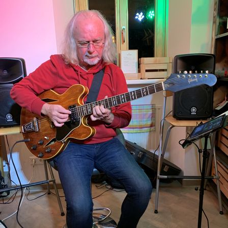 Ein älterer Mann mit langen weißen Haaren und einer Brille spielt Gitarre in einem Raum. Er trägt eine rote Kapuzenjacke und blaue Jeans.