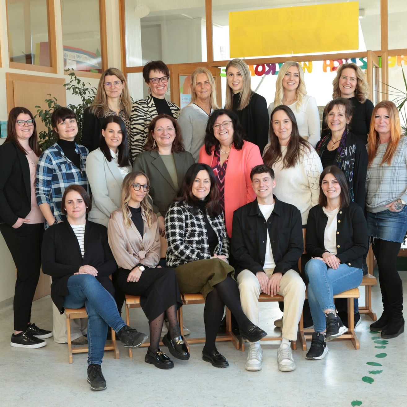 Eine Gruppe von Menschen in formeller und lässiger Kleidung posiert für ein Foto in einem Raum mit Holzbänken und einem gelben Banner.