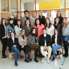 Eine Gruppe von Menschen in formeller und lässiger Kleidung posiert für ein Foto in einem Raum mit Holzbänken und einem gelben Banner.