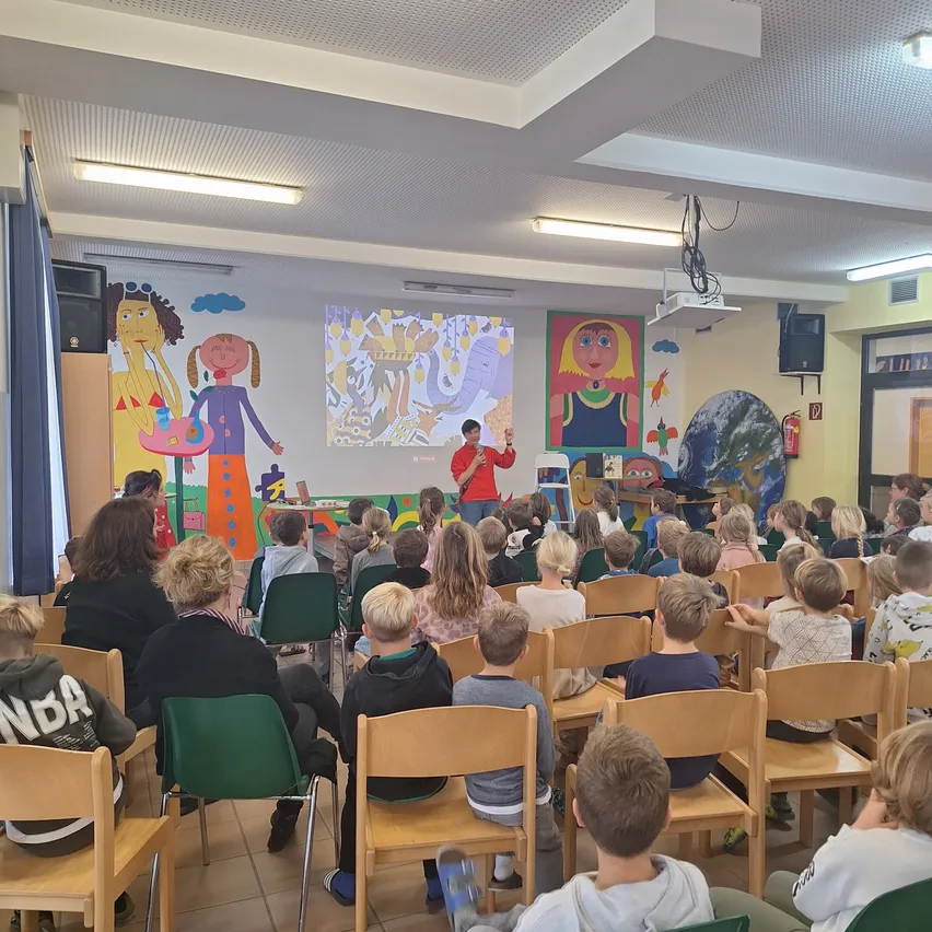 Ein Mann steht vor einem Klassenzimmer mit einer Projektionsleinwand und hält eine Präsentation für einen Raum voller Kinder, die auf Stühlen sitzen. Der Raum hat farbenfrohe Wandgemälde und Deckenleuchten.