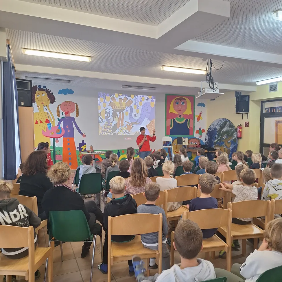 Ein Mann steht vor einem Klassenzimmer mit einer Projektionsleinwand und hält eine Präsentation für einen Raum voller Kinder, die auf Stühlen sitzen. Der Raum hat farbenfrohe Wandgemälde und Deckenleuchten.