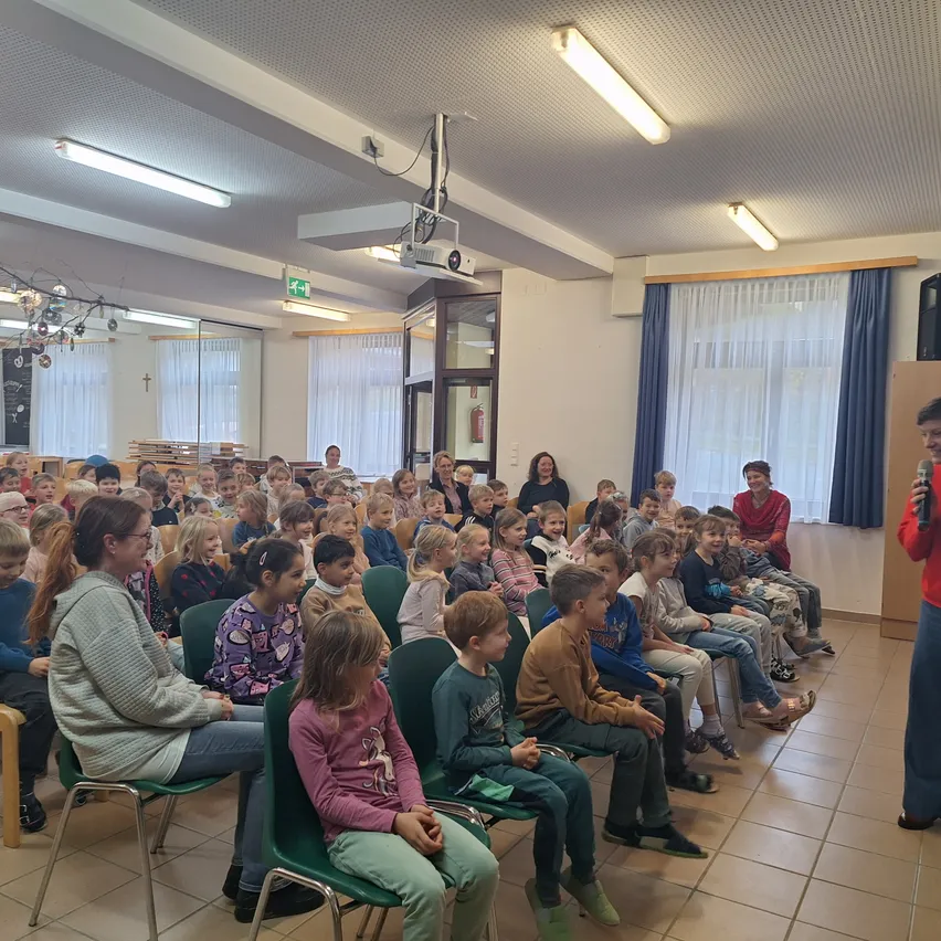 Eine große Gruppe von Kindern und Erwachsenen sitzt in Stühlen vor einem Redner auf der Bühne. Der Raum ist gut beleuchtet mit Deckenleuchten und hat einen Projektor und Vorhänge.