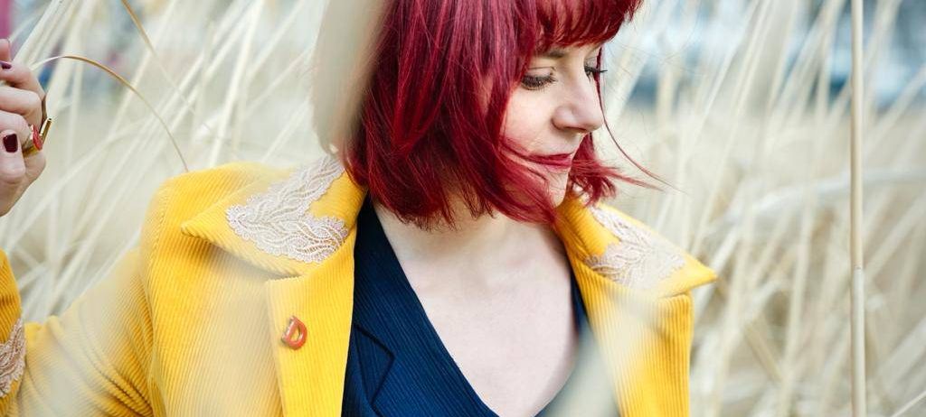 Eine Frau mit roten Haaren und einem gelben Mantel mit weißer Spitzenbordüre, einem blauen Hemd mit weißem Katzendesign, steht lächelnd in einer natürlichen Umgebung.