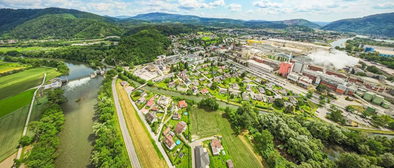 Luftaufnahme einer kleinen Stadt am Fluss, umgeben von grünen Hügeln und Bergen. Rechts ist eine große Fabrik zu sehen.