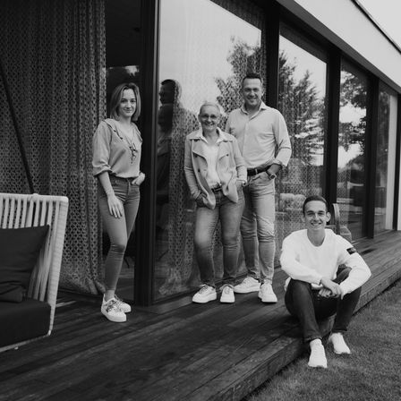 Vier Personen posieren auf einer Veranda eines modernen Hauses. Zwei Frauen stehen hinter zwei Männern. Der Mann auf der rechten Seite trägt eine Uhr und einen Gürtel. Der Mann auf der linken Seite trägt ein Langarmshirt.