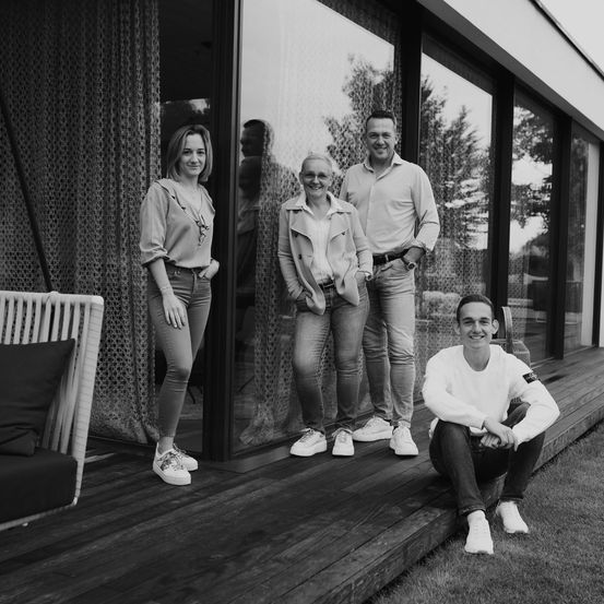 Vier Personen posieren auf einer Veranda eines modernen Hauses. Zwei Frauen stehen hinter zwei Männern. Der Mann auf der rechten Seite trägt eine Uhr und einen Gürtel. Der Mann auf der linken Seite trägt ein Langarmshirt.