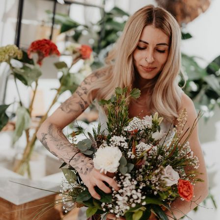 Eine Frau mit Tätowierungen auf den Armen hält einen Blumenstrauß, möglicherweise für eine Hochzeit.