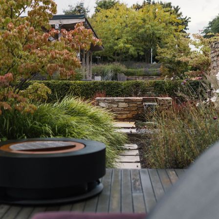Ein gepflegter Garten mit einem Steinpfad, der zu einem kleinen Brunnen führt. Bäume mit roten Blättern und ein hölzerner Pavillon im Hintergrund.