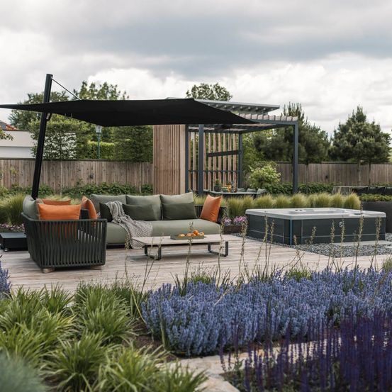Ein moderner Außensitzbereich mit einem Sofa, einem Kaffeetisch und einem Whirlpool unter einem schwarzen Baldachin, einer Holzumzäunung und Grün im Hintergrund.
